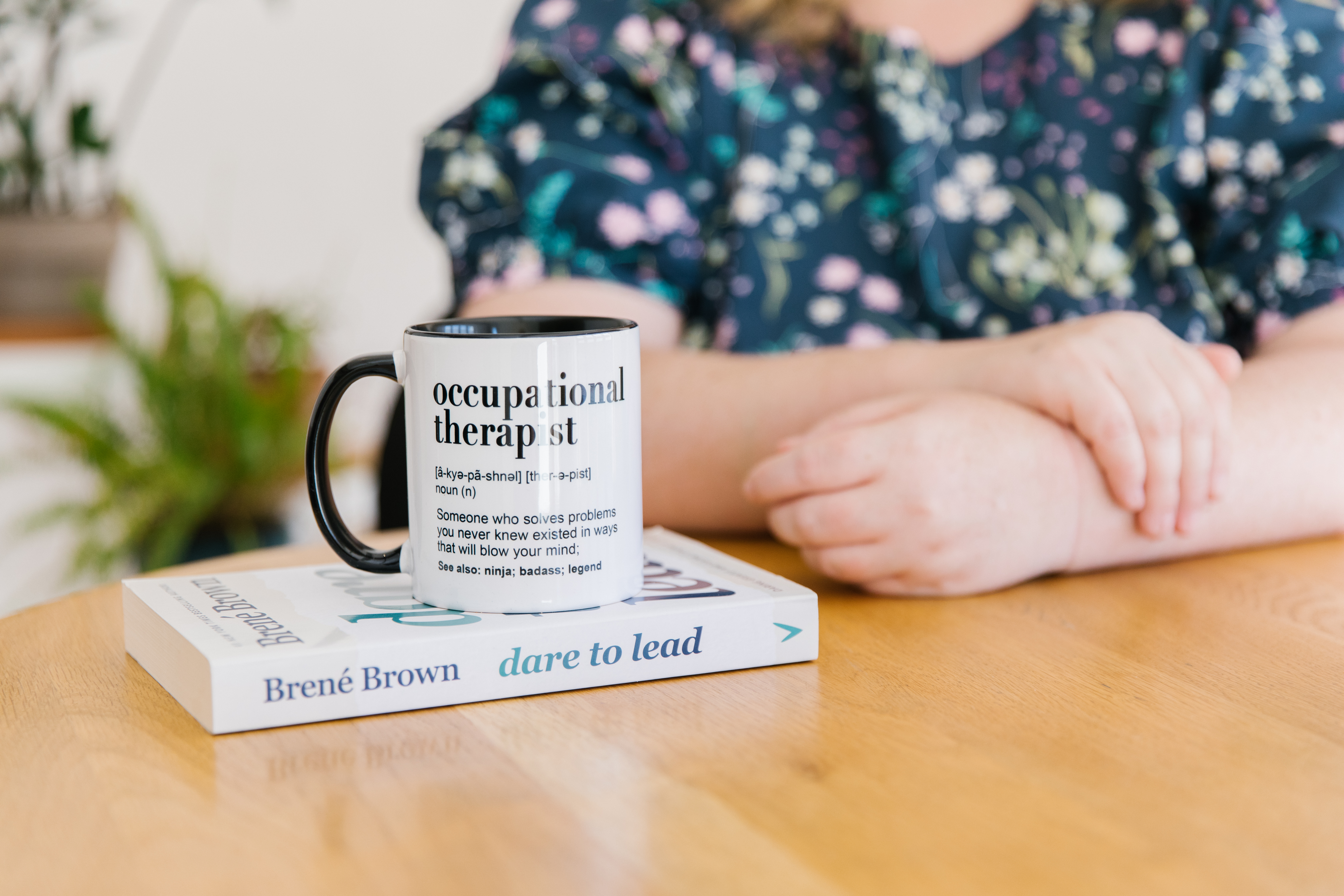 A person sits at a table with their arms crossed, behind a book titled "Dare to Lead" by Brené Brown and a mug labeled "Occupational Therapist.