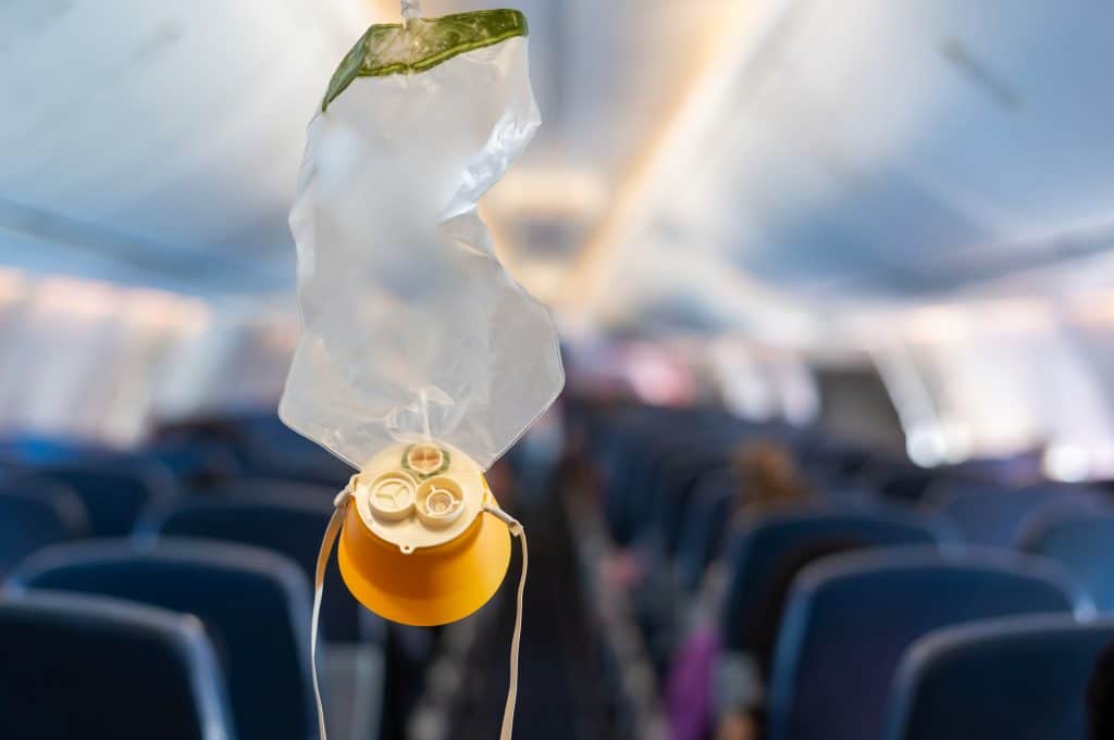 A deployed oxygen mask hanging inside an airplane cabin, with blurred seats and passengers visible in the background.