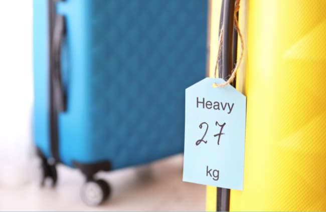 Close-up of a tag reading "Heavy 27 kg" attached to a yellow suitcase, with a blue suitcase visible in the background.