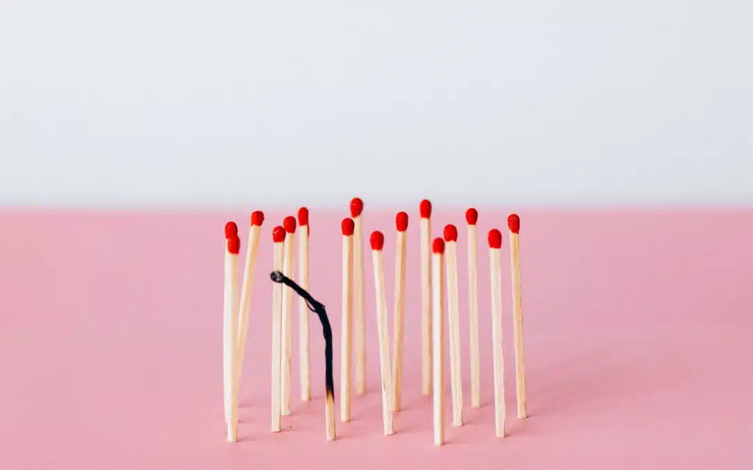 A burnt matchstick stands among unlit matchsticks with red tips against a pink surface and a light gray background.