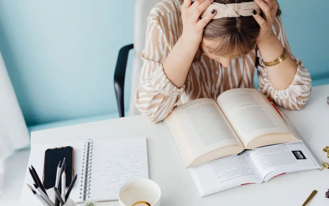 A person holding their head in frustration over an open book on a desk, alongside a notebook, coffee cup, and phone.