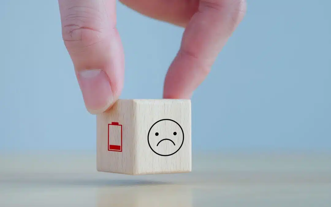 A hand holds a wooden block with a low battery icon and a sad face drawn on it, set against a plain background.